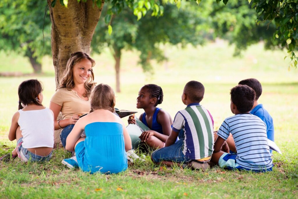 A group of kids and a woman sitting under a tree